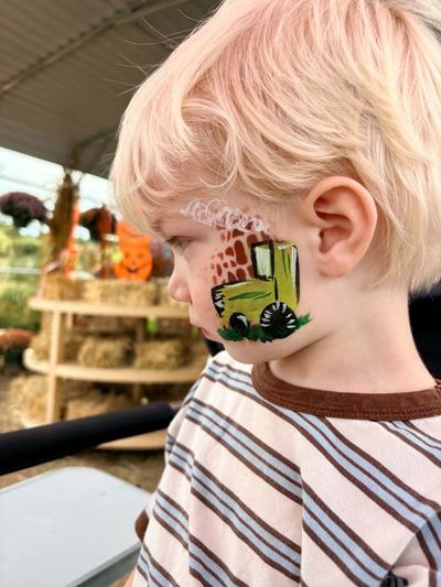 Toddler with a green tractor cheek design at a fall farm, face painting by In His Image, Morristown TN
